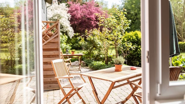 Eine Terrasse im Freien mit Holztisch und -stühlen, einer Holztreppe und Blick auf einen Garten mit verschiedenen Pflanzen und Bäumen.