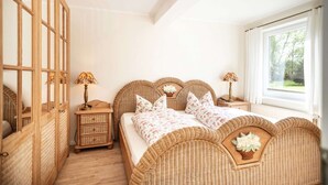 Een slaapkamer met een groot raam, een bed met bloemenbeddelsels, een nachtkastje en een rieten hoofdbord.