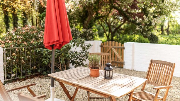 A wooden table with a potted plant and a lantern, accompanied by wooden chairs, set on a gravel surface with a red umbrella in the background.
