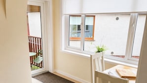 Une pièce comprenant une chaise blanche, une petite table, une fenêtre et une porte donnant sur un espace extérieur avec un banc et des chaises.