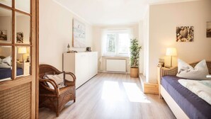 Una habitación de hotel acogedora con cama, una silla de mimbre, una mesita de noche con lámpara, una planta en maceta y vistas al exterior a través de una ventana.
