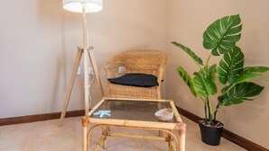 Et hjørneværelse med en fletstol, et lille glasbord, en gulvlampe og en potte med monstera.