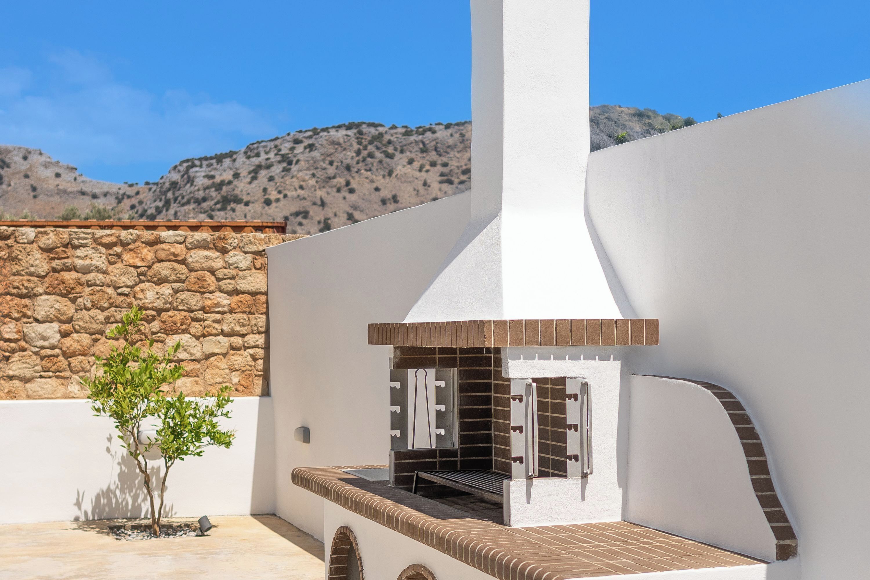 Une terrasse avec un barbecue en briques, un petit arbre et un mur en pierre.