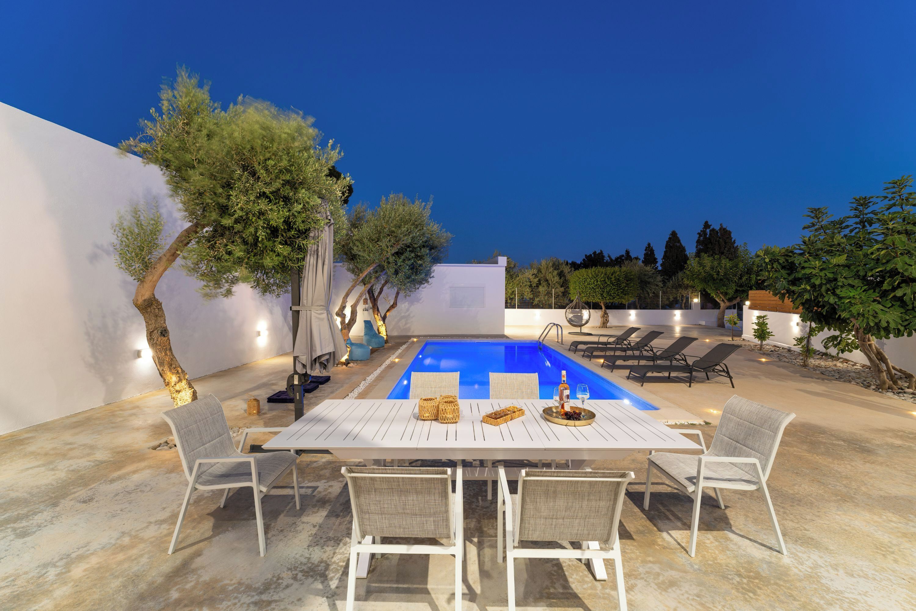 A poolside area with a rectangular pool, white lounge chairs, and a dining table with chairs.