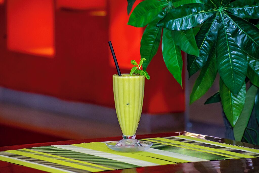 A tall glass of yellow beverage with a black straw and mint garnish on a striped placemat, with a large green plant in the background.