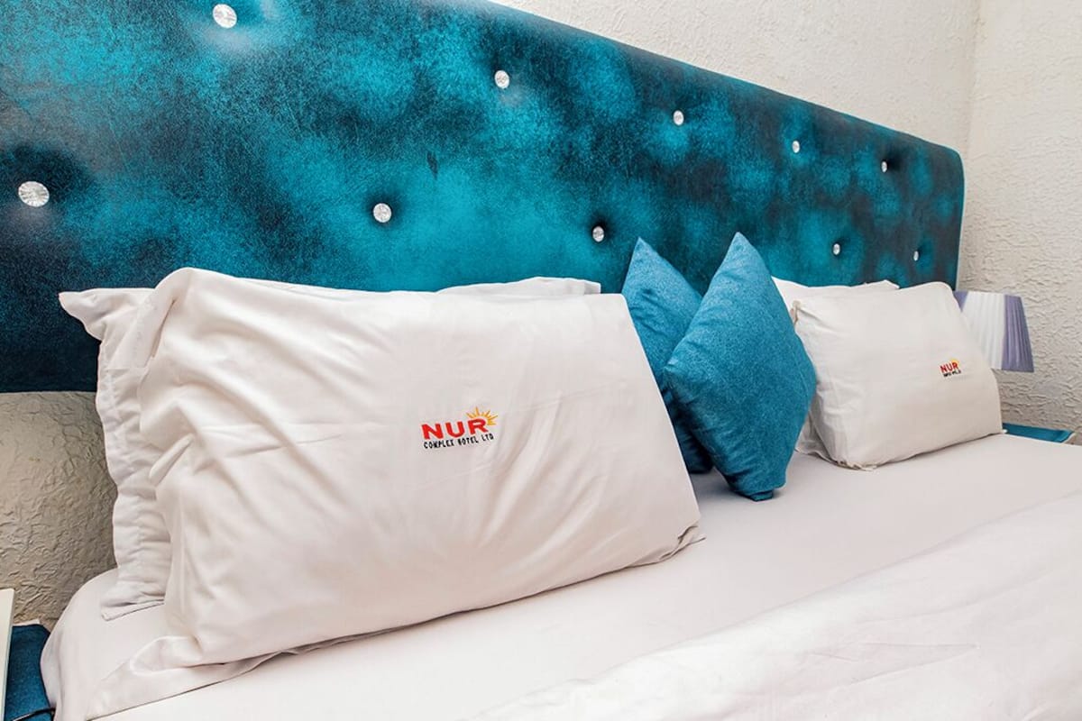 A hotel room with a tufted headboard, white bedding, and pillows with the NUR logo.