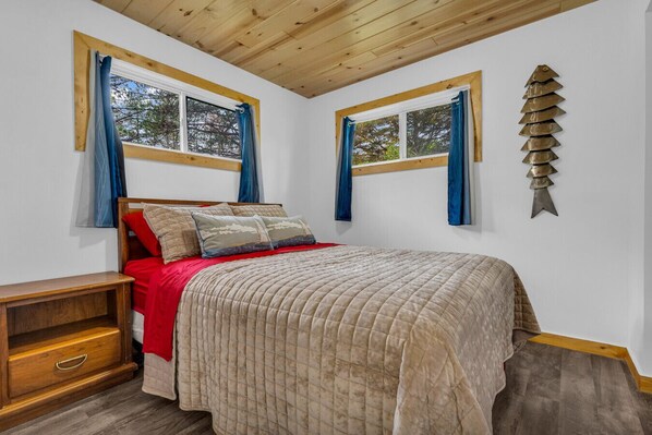 A bedroom with a bed, two windows, a wooden headboard, a bedside table, and a wall-mounted decorative piece. - North Bound 25 Cottage #5 (Lexington)