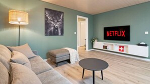 A modern living room with a sofa, ottoman, coffee table, and a mounted TV displaying 'NETFLIX'. - AlpsView Jade/FreeParking/AC/Andermatt/Lucerne (Altdorf)