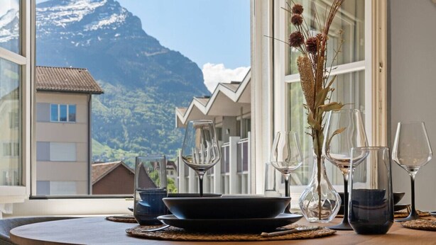 A dining table with wine glasses and a vase of dried plants, set against a mountain view. - AlpsView Jade/FreeParking/AC/Andermatt/Lucerne (Altdorf)