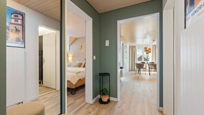 A hallway leading to a bedroom with a bed, a small table, and a potted plant. There is a dining area with a table and chairs visible in the background. - AlpsView Jade/FreeParking/AC/Andermatt/Lucerne (Altdorf)