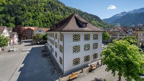 A white building with yellow and black striped windows, surrounded by a cobblestone street and other buildings, with a mountainous backdrop. - AlpsView Jade/FreeParking/AC/Andermatt/Lucerne (Altdorf)