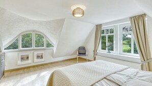 Un dormitorio con una cama grande, una silla, un escritorio con su silla y una ventana con cortinas.