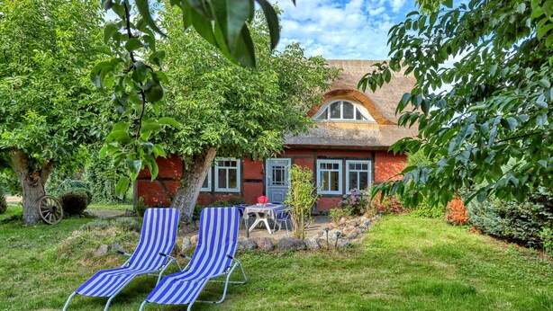 Una casa con fachada de ladrillo rojo, techo de paja y un jardín con dos sillones a rayas y una mesa.