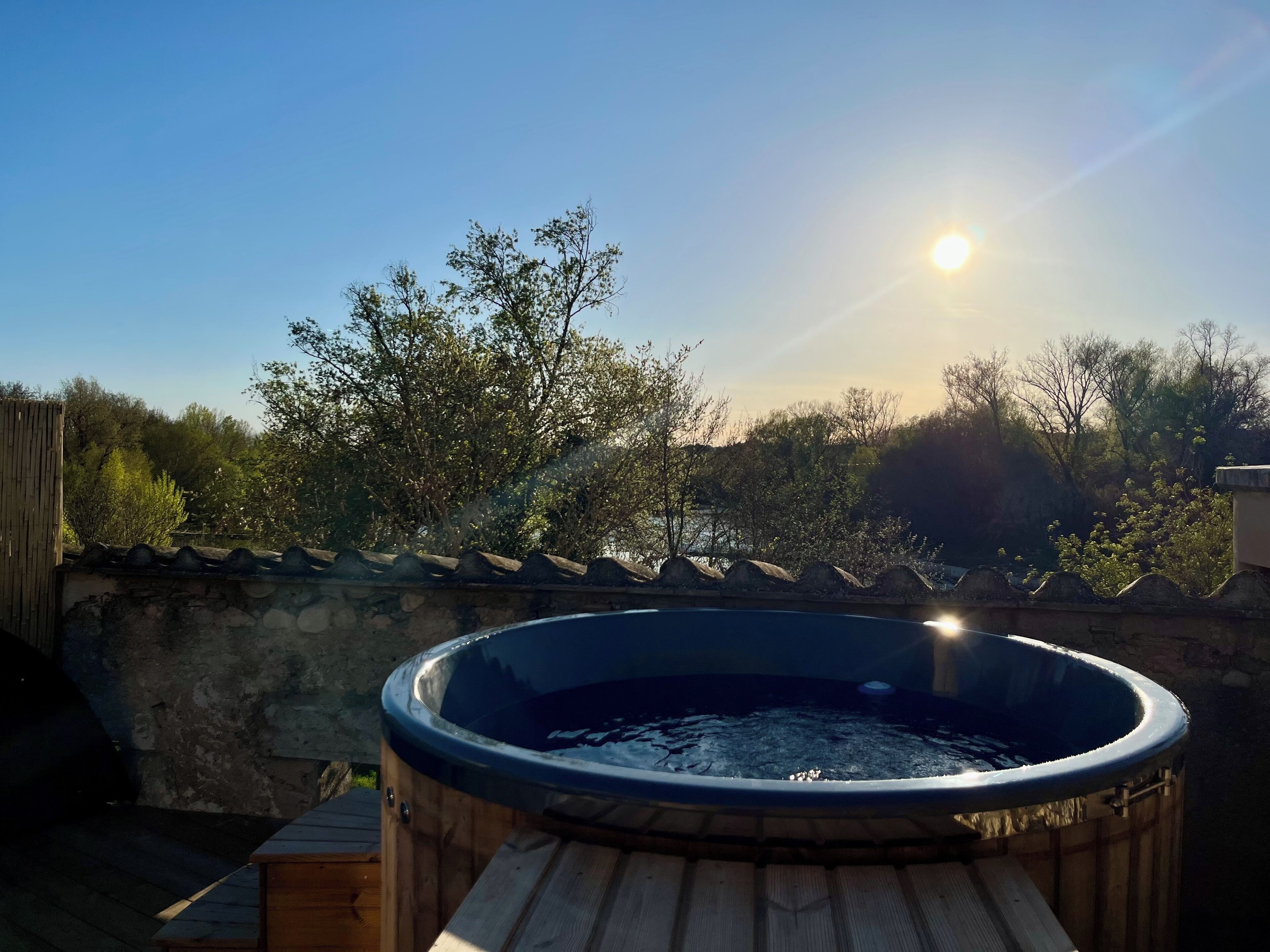 A hot tub on a wooden deck with a stone wall and trees in the background.