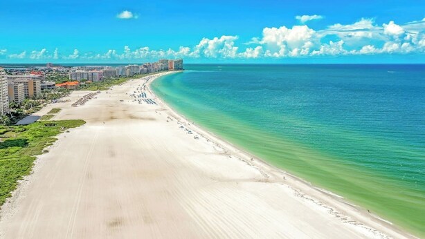 A coastal beach with a wide sandy shore, a clear blue sea, and a row of buildings along the coastline.