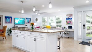 A modern kitchen with white cabinetry, a granite countertop, and a central island. There is a dining area with a table and chairs, a TV mounted on the wall, and artwork on the walls.