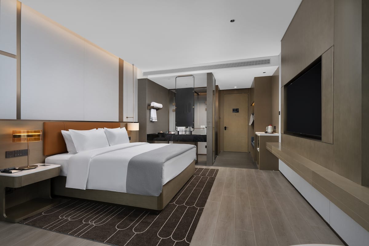 Una habitación de hotel moderna con una cama grande, mesitas de noche, televisor, espejo y kitchenette.