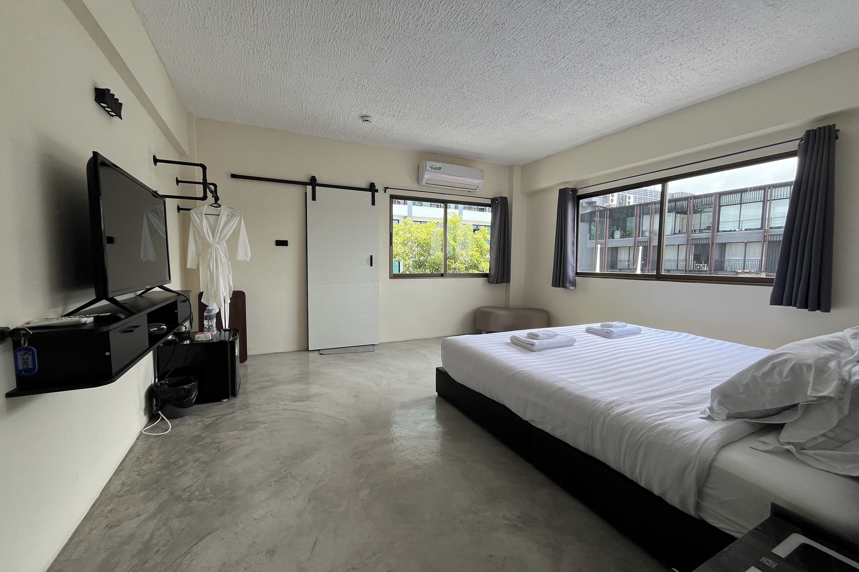 A hotel room with a bed, a TV, a robe, a desk with a computer, and a window with a view of buildings.