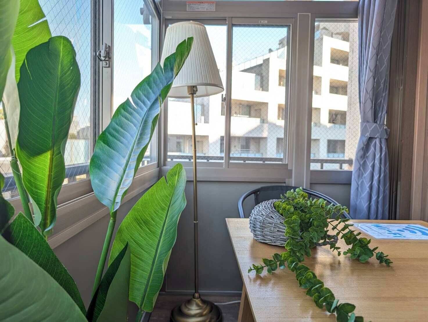 A room with large windows, a floor lamp, a wooden table, and a wicker basket with plants.