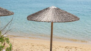 Uma praia com guarda-sóis de palha e espreguiçadeiras, um veleiro ao fundo, e águas calmas.