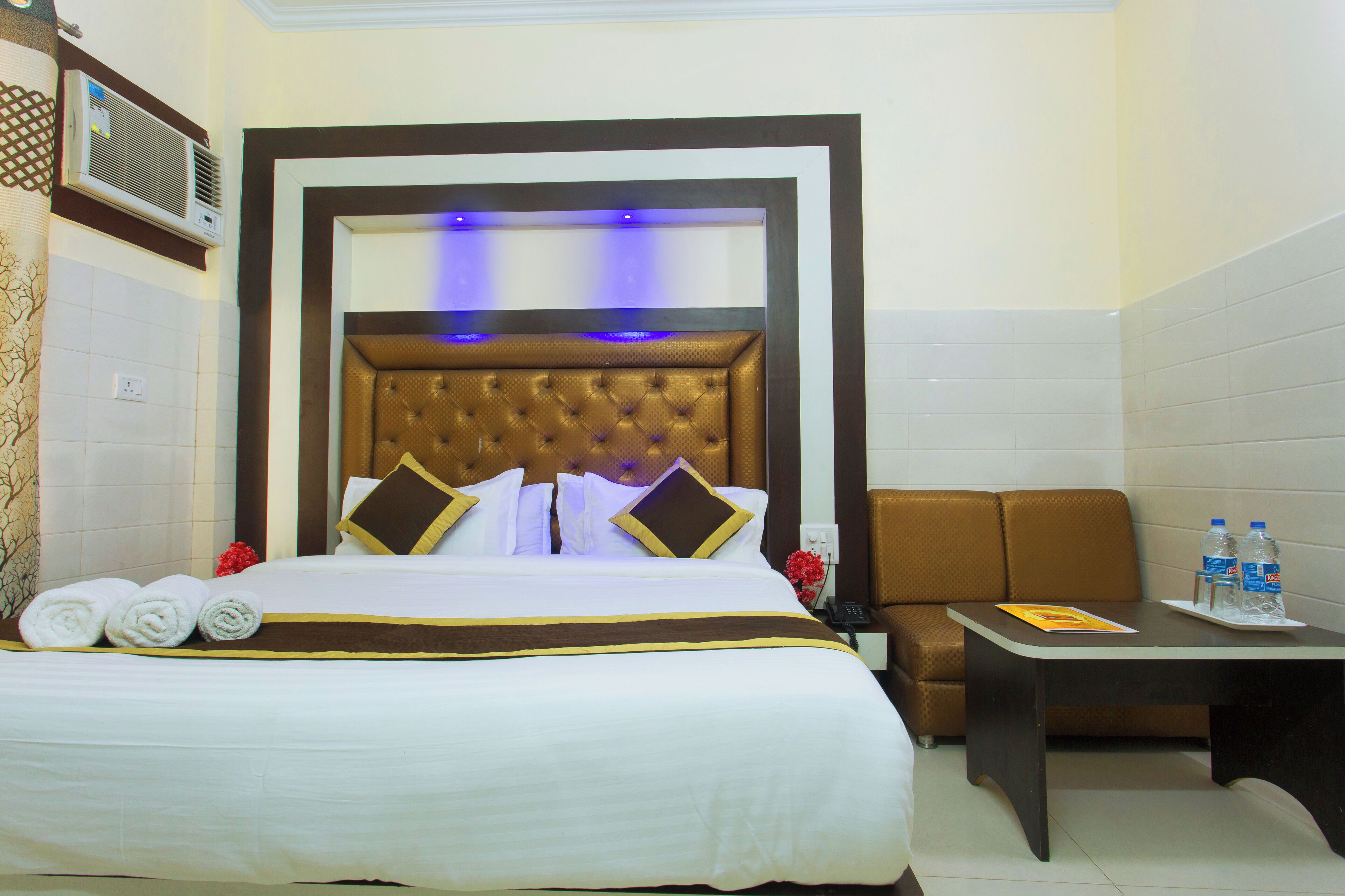 Ein Hotelzimmer mit einem großen Bett, einem mit blauer Beleuchtung versehenen Kopfteil, einer Couch, einem kleinen Tisch mit Wasserflaschen und einer Wand-Klimaanlage.