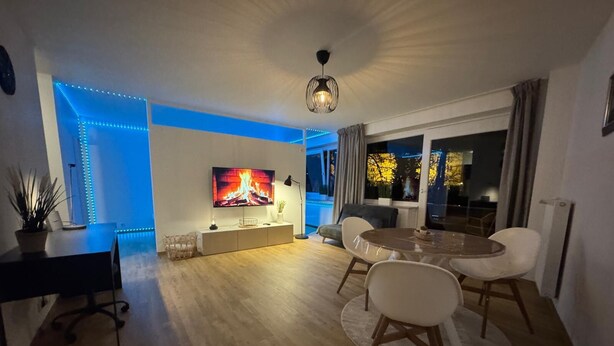 A modern living room with a flat-screen TV, a dining table with chairs, a sofa, and a large window with curtains.