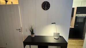 A modern room with a desk, a clock, and a door leading to a kitchen.