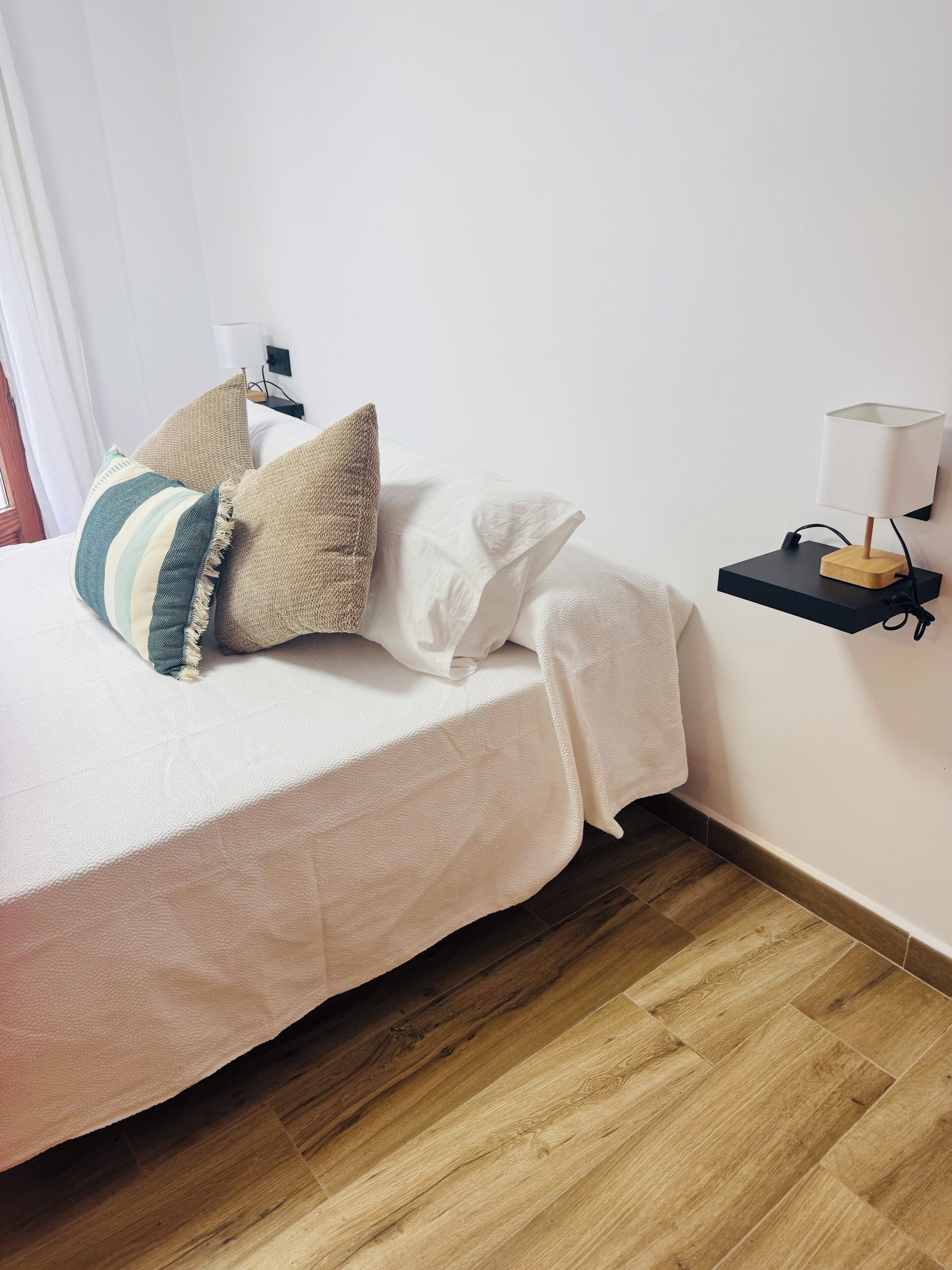 Un lit bien fait, recouvert d’une courtepointe blanche, avec deux oreillers et une table de chevet sur laquelle se trouve une lampe.