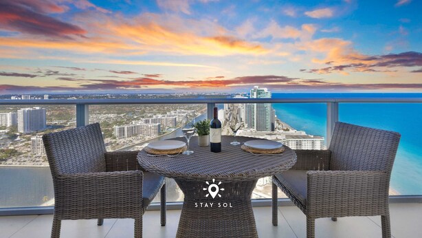 A balcony with wicker furniture, a bottle, and a glass, overlooking a cityscape and the sea. - Stunning 1BR Apt With Ocean View Balcony (Hollywood)