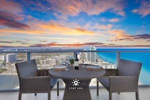 A balcony with wicker furniture, a bottle, and a glass, overlooking a cityscape and the sea. - Stunning 1BR Apt With Ocean View Balcony (Hollywood)