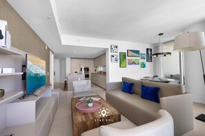 A modern living room with a flat-screen TV, a coffee table, a sofa, and a dining area with a lamp. - Stunning 1BR Apt With Ocean View Balcony (Hollywood)