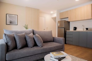 Una sala de estar moderna con un sofá gris, una mesa de centro con un libro y una cocina con armarios de madera y electrodomésticos.