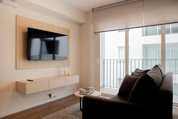 A modern living room with a flat-screen TV mounted on the wall, a sofa, a small side table, and a large window with blinds.