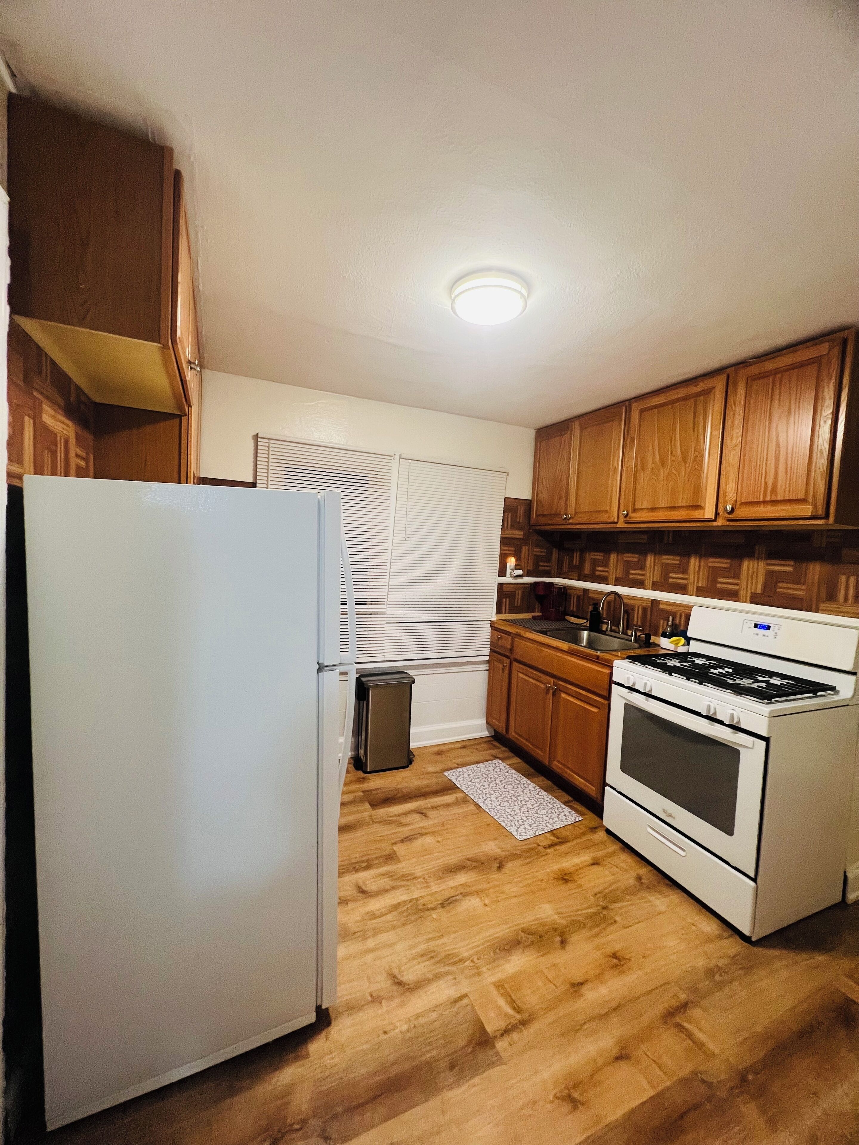 A compact kitchen with wooden cabinets, a white refrigerator, a stove, and a small trash bin.