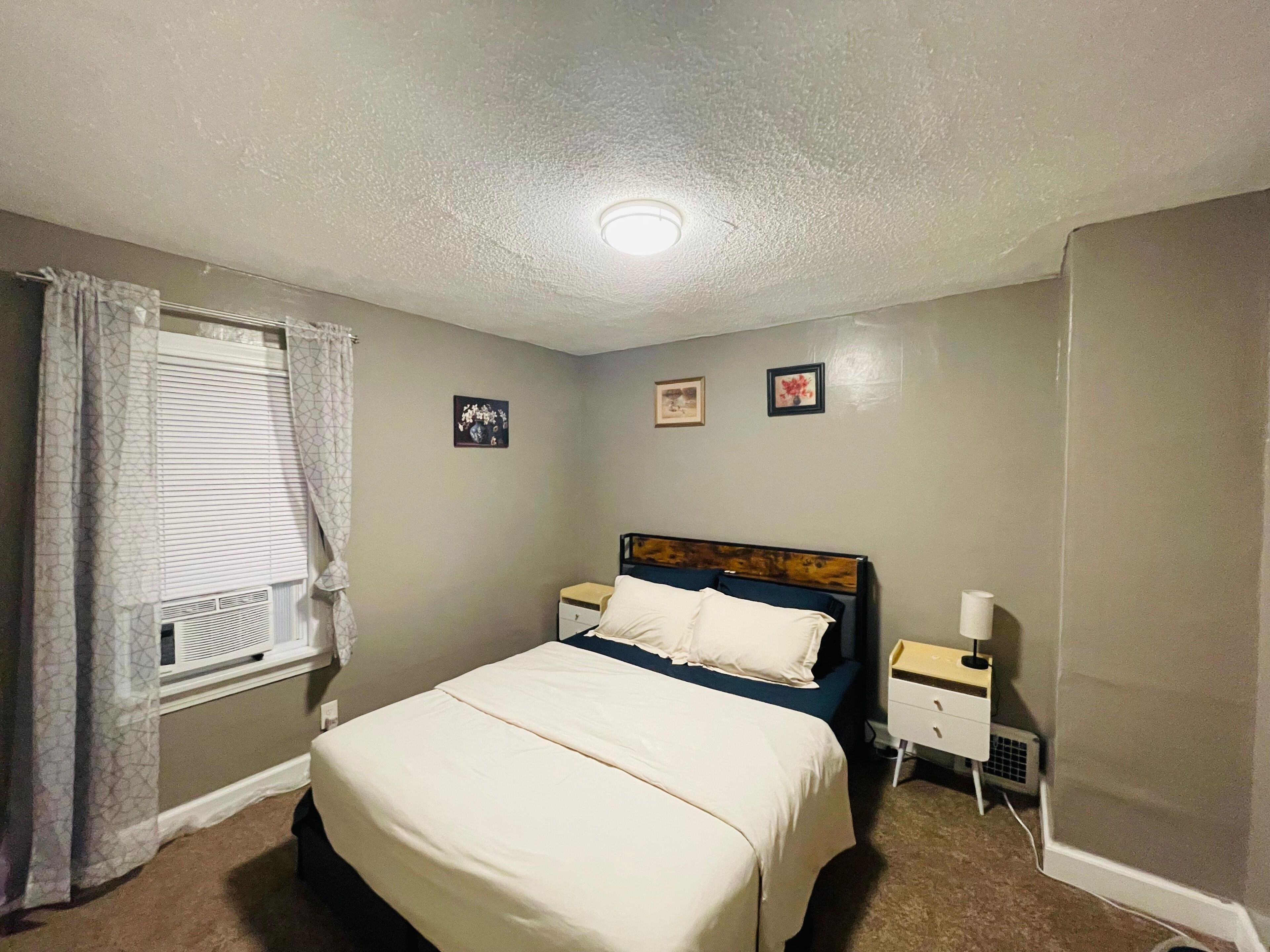 Une chambre à coucher comprenant un lit, une table de chevet, une commode et une fenêtre avec des stores.