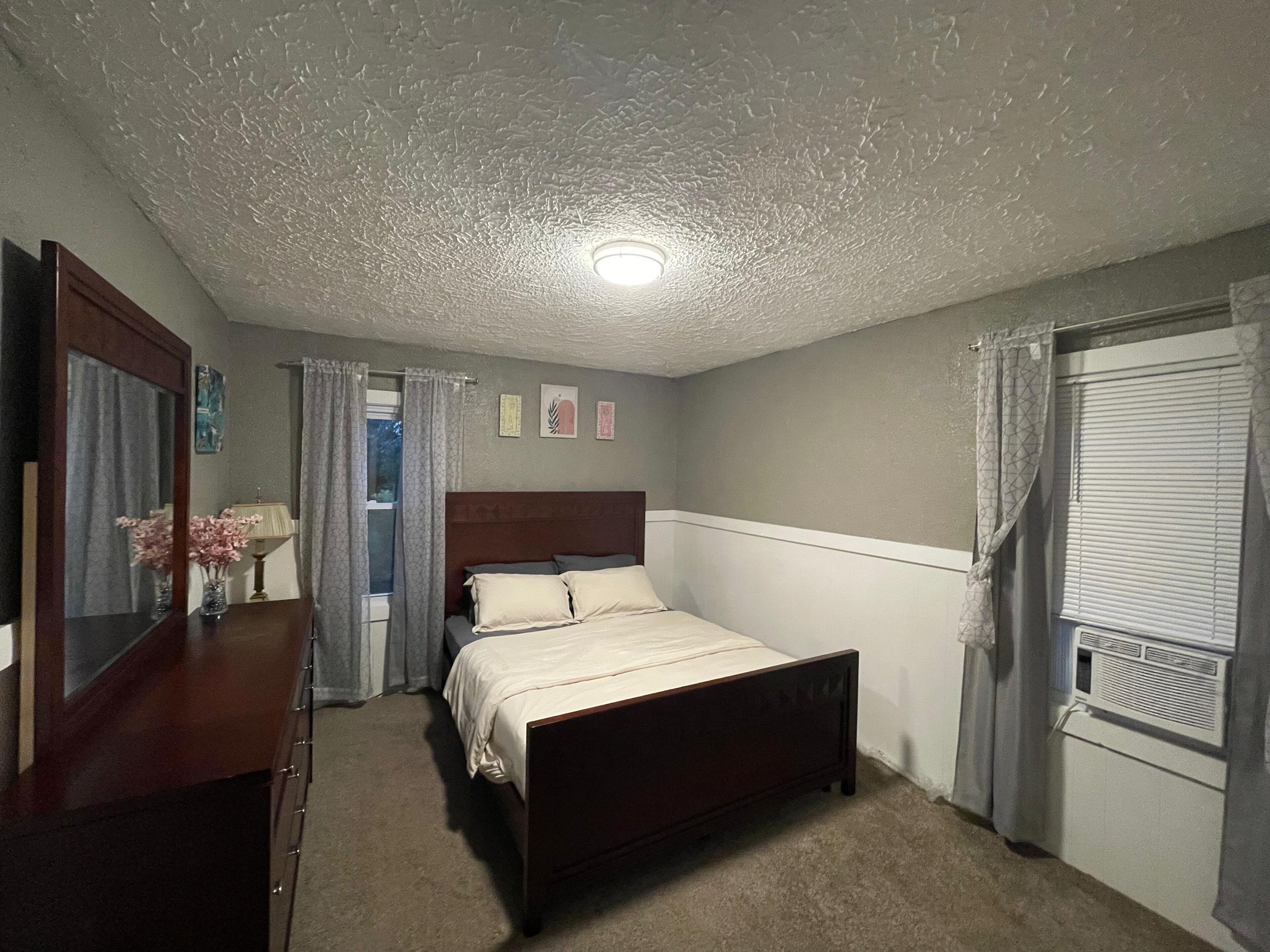 A bedroom with a bed, a dresser, a mirror, a window with curtains, and a ceiling light.