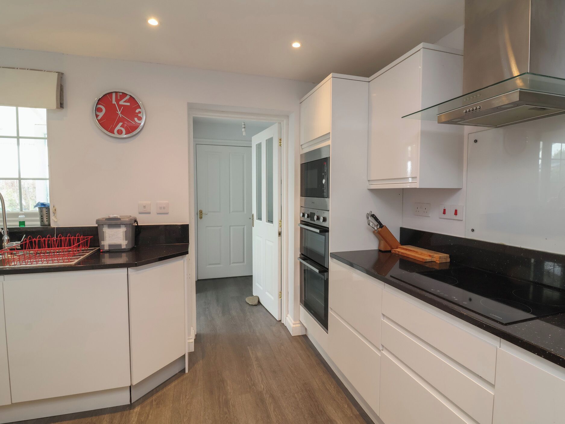 Une cuisine moderne avec des meubles blancs, un plan de travail noir, une hotte en acier inoxydable et une horloge rouge accrochée au mur.