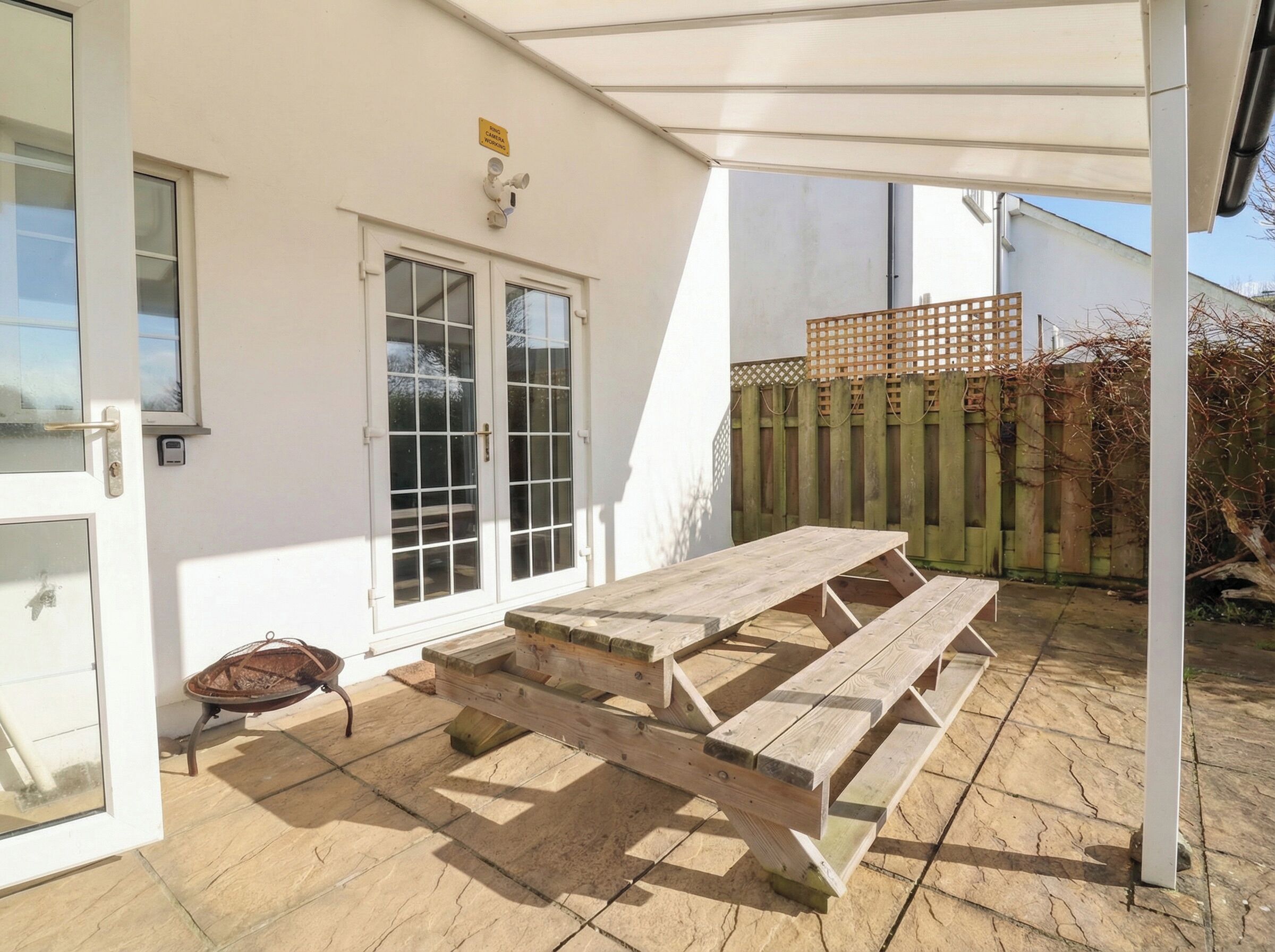 Un patio cubierto con una mesa y bancos de madera, una puerta blanca con paneles de vidrio y una valla de madera al fondo.