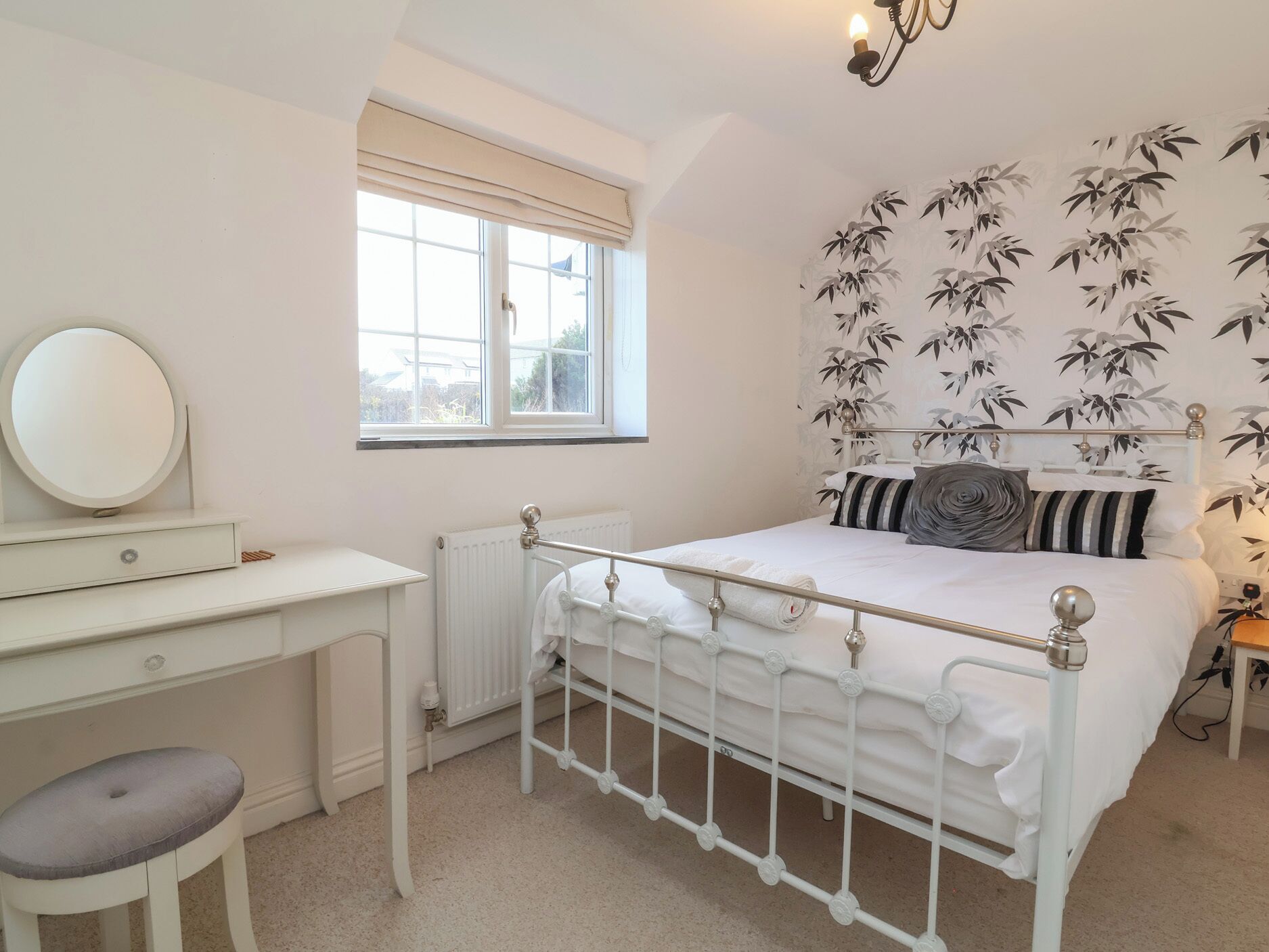 A bedroom with a white metal bed frame, a round mirror, a white desk with a chair, and a window with a view of buildings outside.