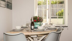 Ein Esstisch für vier Personen, gedeckt mit weißen Tellern, Gläsern und einer Flasche Prosecco. Zwei Stühle, eine Vase mit Blumen und ein Fenster mit Blick auf einen Holzzaun und Grünflächen.