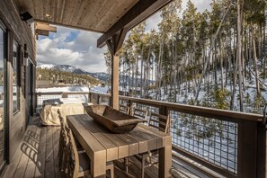 A wooden deck with a table and chairs, overlooking a snowy forest and mountains.