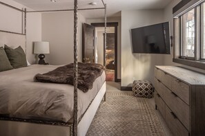 A four-poster bed with a fur throw, a bedside lamp, a TV mounted on the wall, and a patterned rug.