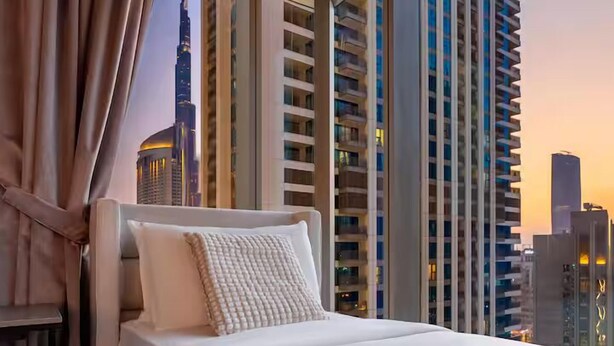 A modern hotel room with a large bed, a view of a city skyline, and a window with a curtain. - Downtown Views (Dubai)
