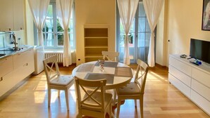 A dining area with a round table, four chairs, and a central vase. There is a kitchen area to the left and a television on a cabinet to the right. - Studio apartment with garden in San Siro (Milano)