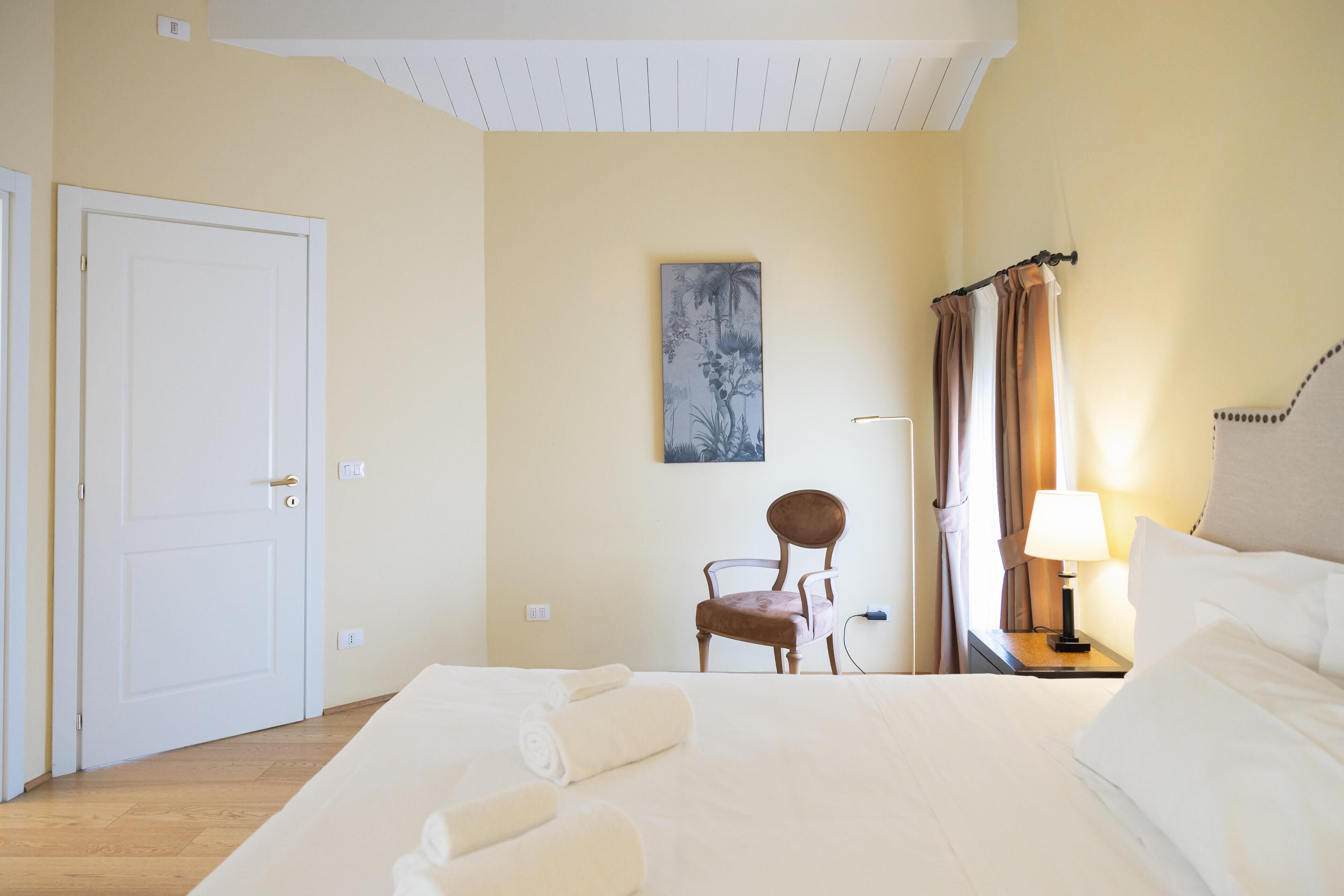 A hotel room with a bed, a chair, a nightstand with a lamp, a painting on the wall, and a door.