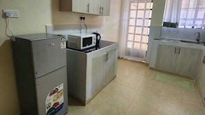A compact kitchen with a refrigerator, microwave, and a small sink. There is a window with curtains and a door leading outside. - Lux Suites Ole Nairi Holiday Homes (Ngong)