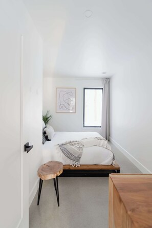 Une chambre à coucher avec un lit, un petit tabouret, une fenêtre et un tableau encadré accroché au mur.
