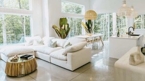 Un salon moderne avec un canapé blanc, une table basse ronde et un coin repas comprenant une table et des chaises blanches. De grandes fenêtres offrent une vue sur la forêt, et il y a également un coin cuisine avec un évier et un plan de travail.