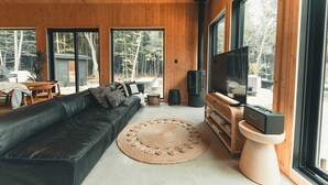 Een moderne woonkamer met een zwarte leren bank, een rond geweven vloerkleed, een televisie op een wit voetstuk en grote ramen met uitzicht op de omgeving.