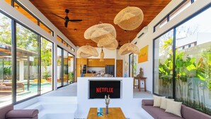A modern living area with a large TV screen displaying 'NETFLIX', a wooden ceiling, and large windows offering a view of the outdoors. - Villa Linaria by Pertama Stay (Ungasan)